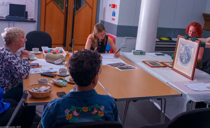A photograph of an arts and crafts event, three women are sat at a table with another women showing them a piece of art.